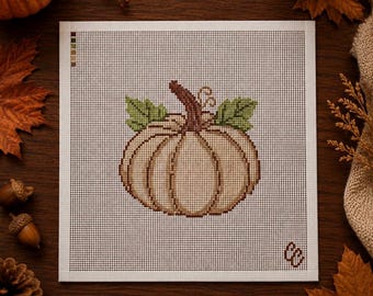 May include: A square needlepoint canvas featuring a cross-stitched design of a light tan pumpkin with a brown stem and two green leaves. The canvas has a white grid background, a small color palette guide in the top left, and initials in the bottom right.