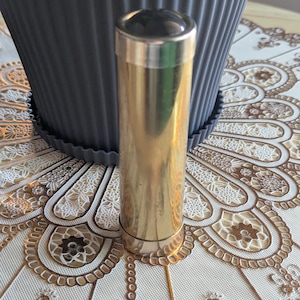 May include: A cylindrical brass kaleidoscope with a polished metallic finish. The object features a smooth, reflective surface and a circular viewing end cap. The design is simple and vertical, showcasing a clean, unadorned exterior body.