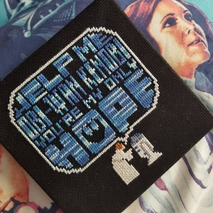 May include: Cross-stitch artwork featuring the Star Wars quote "Help me Obi Wan Kenobi, you're my only hope."  The quote is in blue and white thread on a black background.  Two figures, a white-robed figure and a droid, are stitched in the bottom right corner.