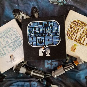 Könnte beinhalten: Drei gerahmte Kreuzstich-Bilder mit Star Wars-Motiven. Das erste Bild ist weiß mit blauem Text, der "Hilf mir Obi-Wan Kenobi du bist meine einzige Hoffnung" lautet. Das zweite Bild ist schwarz mit blauem Text, der "Hilf mir Obi-Wan Kenobi du bist meine einzige Hoffnung" lautet. Das dritte Bild ist weiß mit goldenem Text, der "Wenn jemand hundert Jahre alt ist, erreichst du siehst so gut aus, wirst du nicht".
