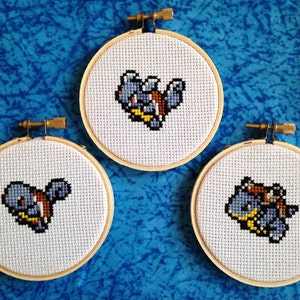 May include: Three embroidery hoops with cross-stitch designs of blue and yellow Pokemon characters. The characters are on white fabric.