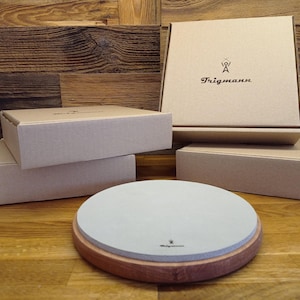 Può includere: Una pietra per affilare circolare grigio chiaro inserita in una base di legno rotonda, posizionata davanti a quattro scatole di cartone beige impilate. La scatola superiore presenta il testo nero "Trigmann" con un logo stilizzato sopra.