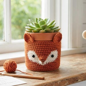 May include: Two terracotta plant pots featuring crocheted covers, one orange fox design and one cream bear design, each containing a green succulent. A small orange yarn ball and a wooden crochet hook sit nearby on the wooden surface.