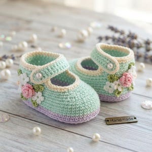 May include: A pair of mint green crochet baby shoes featuring a lavender sole, a single strap with a white pearl button, and decorative floral appliqués in shades of pink, white, and green. A small metal tag labeled Handmade is placed nearby.