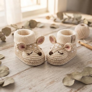 Può includere: Un paio di scarpine per neonati all'uncinetto color beige a forma di cerbiatto, con macchie bianche, piccole orecchie con centro rosa, occhi chiusi ricamati e naso marrone scuro. Le scarpine hanno un bordo a coste color crema e una piccola etichetta.