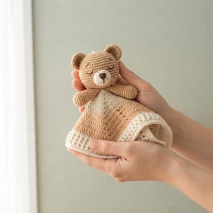 Könnte beinhalten: Eine gehäkelte Schnuffeltuch-Decke mit einem kleinen, hellbraunen Bärenkopf mit gestickten geschlossenen Augen und einer dunkelbraunen Nase. Die angebrachte Decke besteht aus hellbraunem und cremefarbenem Garn mit einem strukturierten Waffelmuster.