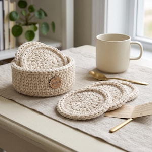 May include: A set of round, cream-coloured crocheted coasters, with several stacked inside a matching circular crocheted basket. The basket features a small, round wooden tag with the text Crafted H&H. The coasters have a textured, woven pattern.