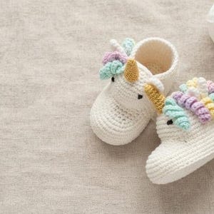 Może przedstawiać: Para szydełkowanych bucików niemowlęcych w kolorze złamanej bieli, stylizowanych na jednorożce. Każdy bucik posiada złoty róg, czarne wyszywane oczy oraz grzywę z pastelowych loków z włóczki w odcieniach różu, żółci, błękitu i zieleni.