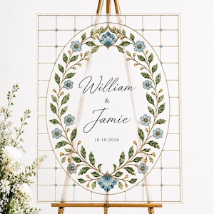 May include: A rectangular clear acrylic wedding sign featuring a stained glass-style grid pattern and a central oval wreath of green leaves and blue flowers. The text reads William & Jamie with the date 10.10.2026 in black script and serif fonts.