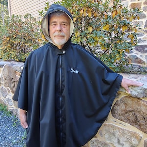 May include: A black hooded poncho featuring a front zipper closure and a light-colored interior lining. The word Scott is embroidered in light blue thread on the upper left chest area of the garment. The poncho has a loose, flowing fit.