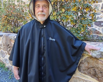 Pode incluir: Um poncho preto com capuz, apresentando um fecho de correr frontal e um forro interior de cor clara. A palavra Scott está bordada com fio azul claro na parte superior esquerda do peito. O poncho tem um corte largo e fluido.