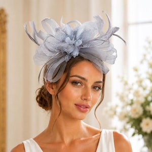 Könnte beinhalten: Ein hellgrauer Fascinator mit geschichteten Netzschleifen, zarten Federn und einem zentralen Blumenakzent. Das Kopfstück ist aus strukturierten, transparenten Stoffbändern gefertigt, die ein voluminöses, mehrdimensionales Erscheinungsbild erzeugen.