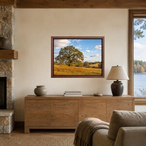 May include: A rectangular landscape wall art print in a dark wood frame featuring a large oak tree standing in a golden grassy field under a blue sky with white clouds. The artwork is displayed on a beige wall above a wooden sideboard cabinet.