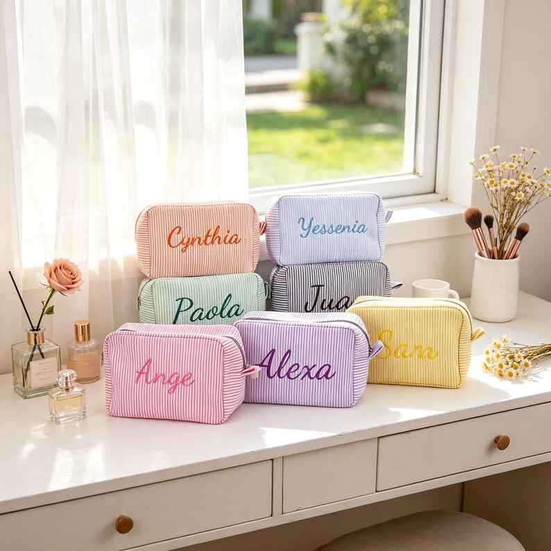 May include: A collection of rectangular zipped pouches made of striped fabric in various pastel colours, each featuring a name embroidered in a cursive font on the front. The pouches are displayed in a group on a white surface.