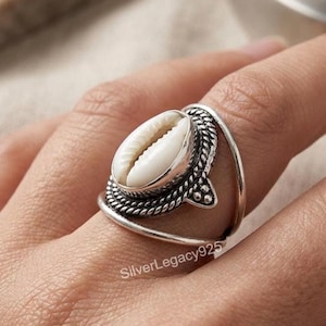 Puede incluir: Un anillo de plata con una concha de cauri blanca central engastada en un marco de metal texturizado y oxidado con un diseño de cuerda trenzada y una banda abierta lisa. El texto SilverLegacy925 es visible en la parte inferior del anillo.