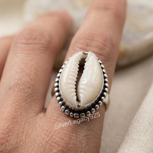 Puede incluir: Un anillo que presenta una concha de cauri blanca central engastada en una banda de plata pulida. El engaste está enmarcado por un borde decorativo de plata con cuentas. El texto SilverLegacy925 es visible en la parte inferior del anillo.