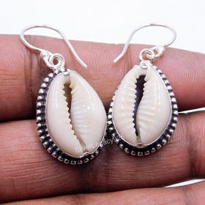 Può includere: Un paio di orecchini pendenti con conchiglie di ciprea bianche naturali incastonate in montature d'argento lucido con bordo perlato. Gli orecchini pendono da ganci d'argento e riportano il testo SilverLegacy925 visibile sulle conchiglie.