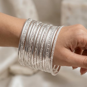 Puede incluir: Un conjunto de diez brazaletes de plata con diversas texturas y patrones, incluyendo grabados florales, líneas onduladas y un diseño trenzado. Las pulseras están pulidas con un alto brillo, creando una superficie metálica reflectante.