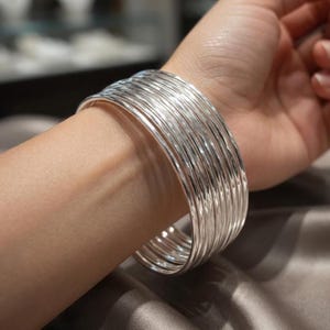 Può includere: Un set di molteplici bracciali rigidi sottili color argento lucido indossati insieme al polso. I bracciali presentano una finitura metallica liscia e riflettente e una forma circolare, creando un aspetto sovrapposto attorno al braccio.