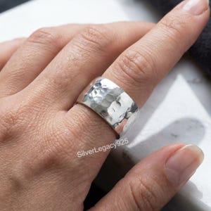 Può includere: Un ampio anello in argento caratterizzato da una superficie martellata e testurizzata con finitura lucida. La fascia ha una larghezza considerevole e un aspetto metallico riflettente. Il testo SilverLegacy925 è visibile sulla superficie dell'anello.