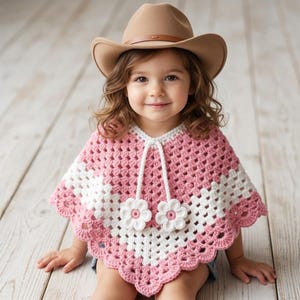 May include: A tan felt cowboy hat and a pink and white crocheted poncho. The poncho features a granny square pattern, a scalloped hem, and two white crocheted flower embellishments attached to long white cords hanging from the neckline.