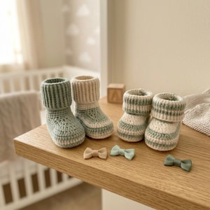 May include: Two pairs of handmade crochet booties in sage green and cream. One pair features a solid sage green ribbed cuff with a cream base, while the other pair has horizontal stripes in sage green and cream. Three small ribbon bows are included.
