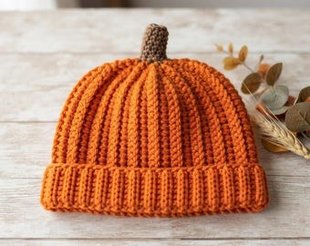 May include: A bright orange crocheted beanie hat with a ribbed texture and a small brown crocheted stem at the top. The hat features a thick folded brim and is constructed from yarn, mimicking the appearance of a pumpkin shape.