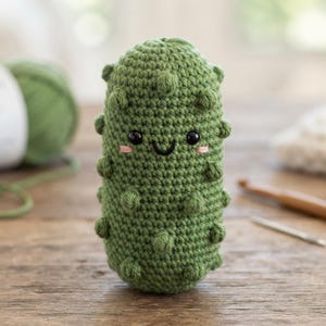 May include: A small, cylindrical, green crochet pickle plush toy featuring a textured surface with raised bumps, two black plastic bead eyes, a stitched black smile, and two small pink embroidered lines representing rosy cheeks.
