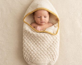 May include: A cream-colored crocheted baby cocoon with a hood, featuring a textured stitch pattern and a mustard yellow trim along the edge. The cocoon is secured with three small wooden buttons on the side, enclosing a newborn infant inside.