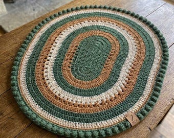 May include: An oval crochet placemat featuring concentric rings of forest green, cream, and tan yarn. The border is finished with a decorative bobble stitch edge. A small, rectangular brown leather tag with the embossed text MCA is attached.