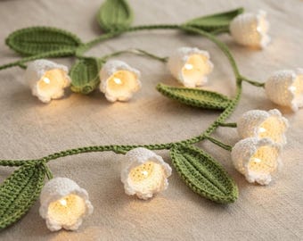 May include: A string of decorative lights featuring crocheted white lily of the valley flowers and green crocheted leaves. Each white bell-shaped flower contains a small glowing light bulb, connected by a green crocheted vine structure.