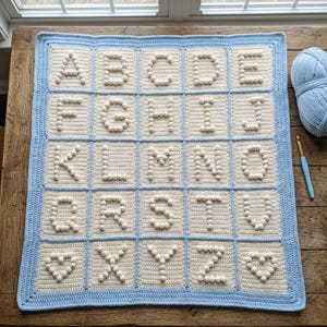 May include: A rectangular crochet blanket featuring a grid of cream-colored squares, each containing a raised bobble-stitch letter of the alphabet from A to Z, with two heart shapes, all bordered by a light blue frame and matching blue trim.
