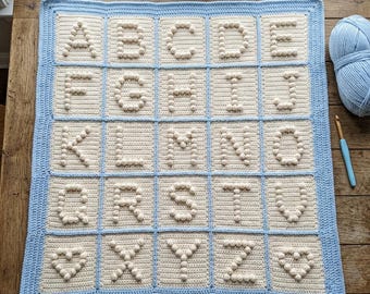 May include: A rectangular crochet blanket featuring a grid of cream-colored squares, each containing a raised bobble-stitch letter of the alphabet from A to Z, with two heart symbols. The blanket has a light blue border and grid lines.