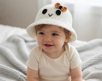 May include: A white crocheted bucket hat featuring a ghost-like face with two black circular eyes and a small black V-shaped mouth. The hat is adorned with a small, textured brown and black bow detail positioned on the upper right side.