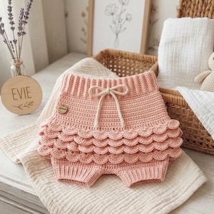 May include: A pair of light pink crochet bloomers featuring a high-waisted design with a drawstring tie and three rows of scalloped ruffles. The garment is crafted with a textured knit pattern and includes a small circular wooden tag on the side.
