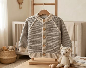 May include: A long-sleeved, textured knit cardigan in a light gray color with cream-colored ribbed trim at the collar, cuffs, and hem. The cardigan features five round wooden buttons down the center front and is displayed on a wooden hanger.