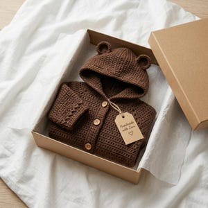 May include: A dark brown crocheted hooded cardigan with bear ears on the hood, featuring three light-colored wooden buttons down the front. A beige paper tag attached to the cardigan with twine displays the text Handmade with Love and a heart.