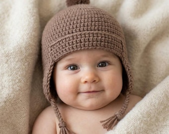May include: A brown crocheted hat featuring a textured grid pattern, a small pom-pom on the crown, and two braided earflaps that extend downward, each finished with a tassel at the end. The hat is crafted from yarn in a solid, earthy brown tone.