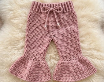 May include: Dusty rose crochet pants featuring a ribbed elasticized waistband with a functional drawstring tie. The pants have a flared leg design with wide, ruffled hems. The crochet stitch pattern is consistent throughout the garment.