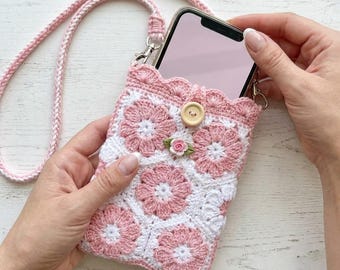 May include: A crochet phone pouch featuring a pink and white floral granny square pattern with a scalloped top edge. The pouch includes a long braided pink strap, a single wooden button closure, and a small decorative fabric rose embellishment.