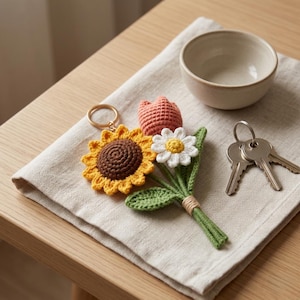 May include: A crochet keychain featuring a bouquet of three flowers: a yellow sunflower with a brown center, a white daisy with a yellow center, and a pink tulip. The stems are green, tied together with light brown yarn, and attached to a gold ring.