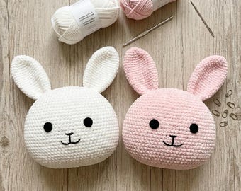 May include: Two crocheted plush rabbit heads, one white and one light pink, each featuring black circular eyes and a stitched black mouth. Included are two skeins of matching yarn, a silver crochet hook, and several small metal stitch markers.