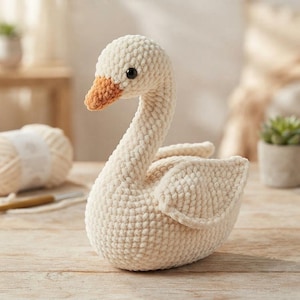 May include: A cream-colored crocheted swan plush toy with a textured, soft yarn finish. The swan features a small, orange-brown beak and a single black bead eye. The body is rounded with detailed wing stitching, capturing a swan in a resting pose.