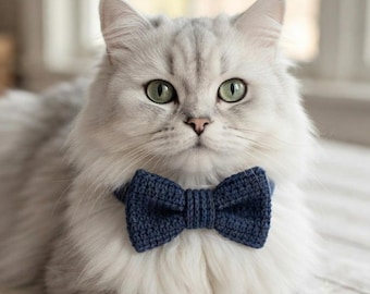 May include: A long-haired silver cat with green eyes wearing a dark blue crocheted bow tie. The bow tie features a textured, hand-stitched appearance with a central knot detail, secured around the neck of the cat with a matching blue band.