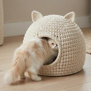 Op de afbeelding: Een crèmekleurig, koepelvormig kattenbed gemaakt van dik, grof gehaakt garen. Het bed heeft twee kleine, puntige oren aan de bovenkant en een grote, ronde opening aan de voorzijde. Een langharige, lichtgekleurde kat zit gedeeltelijk in het bed.