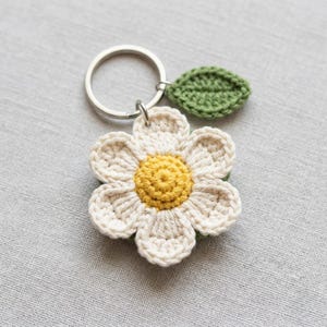 May include: A crochet keychain featuring a white flower with a yellow center and a small green leaf attached to a silver metal ring. The flower has eight rounded petals and a textured, circular yellow core, while the leaf is oval-shaped.