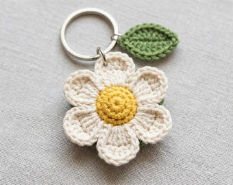 Op de afbeelding: Een gehaakte sleutelhanger in de vorm van een witte bloem met een geel hartje en een klein groen blaadje, bevestigd aan een zilverkleurige metalen ring. De bloem heeft acht afgeronde bloemblaadjes en een textuurrijk, rond geel centrum.