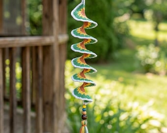 May include: A spiral-shaped crochet hanging ornament featuring a repeating pattern of green, yellow, and brown yarn with a white border. The ornament is suspended from a metal hook and finished with a beaded accent and a multi-colored yarn tassel.