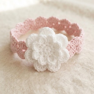 Può includere: Una fascia all'uncinetto rosa chiaro con un bordo smerlato e un grande fiore all'uncinetto bianco a più strati fissato al centro. L'accessorio è realizzato in filato con una trama a maglia visibile su tutto il pezzo.