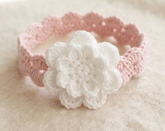 Op de afbeelding: Een lichtroze gehaakte hoofdband met een geschulpte rand en een grote, gelaagde witte gehaakte bloem in het midden. Het accessoire is gemaakt van garen met een zichtbare steekstructuur over het gehele item.
